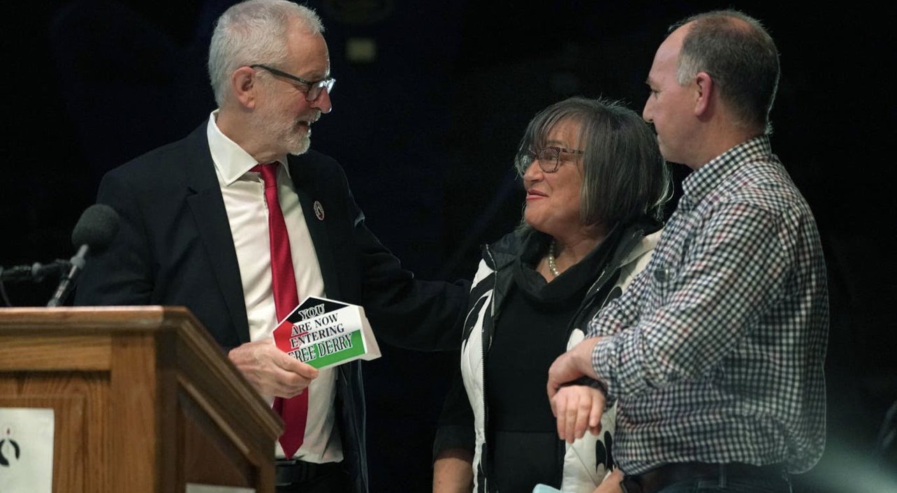 Jeremy Corbyn standing next to a podium, holding a "You are Now Entering Free Derry" sign and speaking to a woman standing next to a man